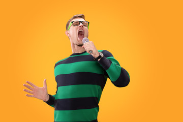 Young man in glasses singing song in microphone. Young posing isolated on yellow orange background....