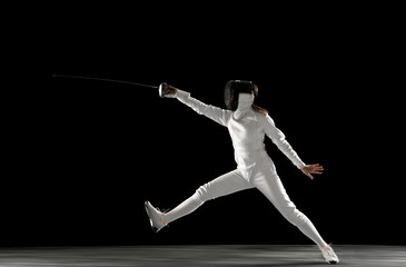 Targets. Teen girl in fencing costume with sword in hand isolated on black background. Young female model practicing and training in motion, action. Copyspace. Sport, youth, healthy lifestyle.