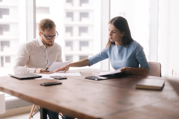 Two colleagues are studying the working documents of the new business project at workplace in the light modern office room against large window. Concept of office life.