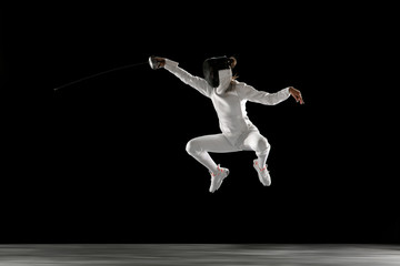 Catching moment. Teen girl in fencing costume with sword in hand isolated on black background....
