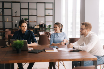 Young business man tells his colleagues about the project in modern office meeting against large panoramic window. Young man and woman listen to the speaker. Concept of office life.