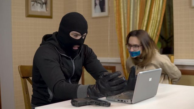 A burglar is trying to break into a laptop by taking a young woman hostage. A masked thug sits in a house and cannot break into a laptop and threatens a hostage with a gun. Theft of data from a