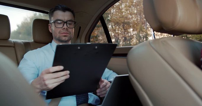 Caucasian attracive businessman in glasses working at the back seat of the car while going to office - checking documents in folder and typing on the laptop computer.