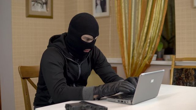 A robber is trying to hack into a laptop. A masked thug is sitting in a house and cannot hack into a laptop. Theft of data from a computer.
