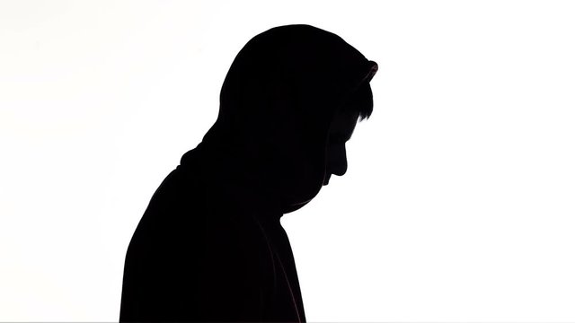 silhouette of a young man puts on a hood of a sweatshirt and goes on white isolated background, unknown person, guy hides face, concept fashion