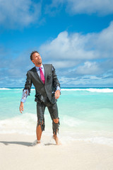 Castaway businessman emerging from tropical sea onto the beach with shredded suit and exhausted...