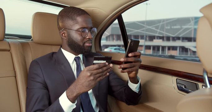 African American young handsome businessman in glasses, suit and tie shopping online with a credit card on the smartphone while sitting on he back seat of expensive car.