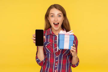 Amazed shocked excited ginger girl in shirt standing with wrapped gift box and mobile phone in her hands, holding present, looking with surprised face. indoor studio shot isolated on yellow background