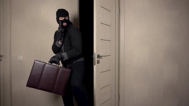 The robber climbed into the house. The masked bandit carefully entered through the door to the room. The robber held a suitcase and a gun in his hands.