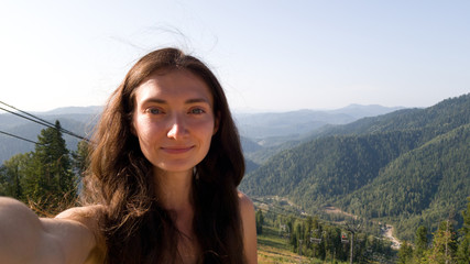 A brunette girl with long hair takes a selfie on the background of beautiful green hills. The forest and the horizon. Cable car in the background