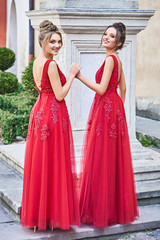 Two beautiful bridesmaids girls blonde and brunette ladies wearing elegant full length red chiffon bridesmaid dress with lace and holding flower bouquets. European old town location for wedding day.