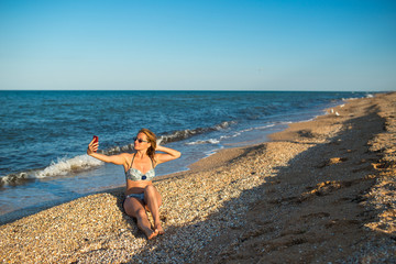 Fototapeta na wymiar Beautiful young woman in a swimsuit makes selfie on a smartphone while relaxing on the beach by the sea on a sunny warm summer day. Social networking and leisure concept. Copyspace