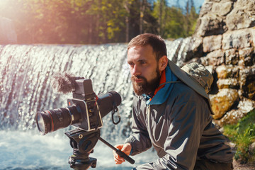 Travel photographer bearded man close-up with professional film camera on tripod shooting mountain...