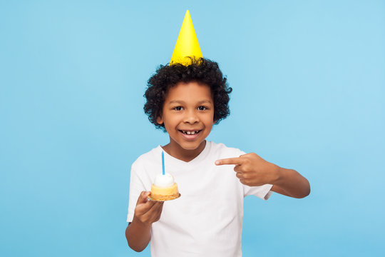 Little Adorable Happy Boy With Funny Party Cone On Head Pointing At Cake With Candle And Looking At Camera With Toothy Smile, Expressing Pure Child Happiness. Studio Shot Isolated On Blue Background