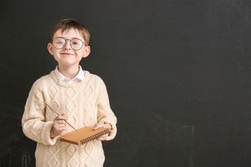 Little schoolboy with notebook near blackboard in classroom