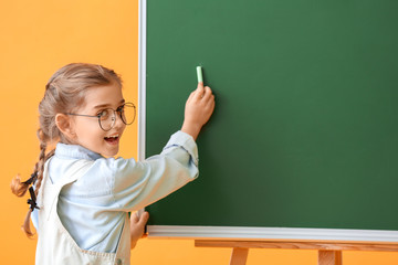 Little schoolgirl writing on blackboard against color background