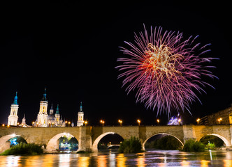 Fototapeta premium colorful fireworks exploding in the dark night sky creating light and color effects above the bridge and close to Basilica of Pilar. Light from fireworks and streetlights reflecting on river water