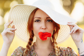 Portrait of beautiful redhead woman in white hat on green field with poppy flower in her mouth