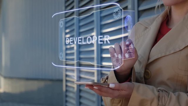 Unrecognizable businesswoman interacts HUD hologram with text Developer. Woman in the coat uses the technology of the future mobile screen on the background of the city