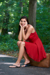 Sexy brunette woman in short red dress waits on side of the road sitting on vintage suitcase - head in hands