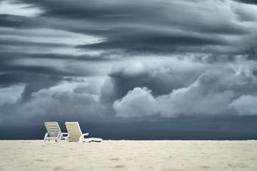 Two empty sun beds standing on white sand beach over a big gray stormy clouds as background.