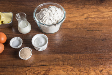 White flour in a glass bowl container, dry yeast, salt, and sugar in small ramekin. 3 pieces of brown chicken eggs, butter, and milk in small glass jar on wood table surface.