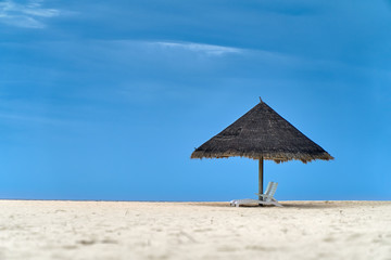 Pair of empty sun beds in shade of straw umbrella on white sand beach. Clear deep blue sky on background