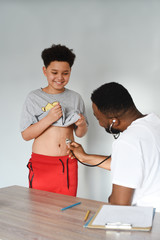 An african american doctor with his mixed preteen male. A cute black paediatrician examining his young mixed preteen boy patient with a stethoscope.