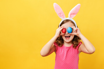 Cheerful happy child in the ears of a bunny holds two Easter eggs in his eyes. Portrait of a little girl on a yellow background in the studio