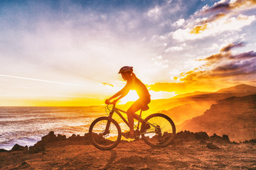 Mountain biking cyclist girl riding MTB bike on coast trail against sunset. Silhouetter of woman...