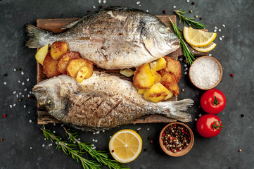 grilled dorado fish and fried potatoes served on a cutting board with spices and lemon on a stone background