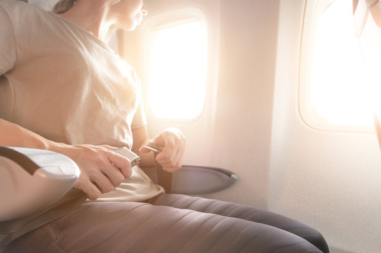 Close-up Of A Young Woman Fastens A Seat Belt While Sitting In A Passenger Airplane Chair By The Window. The Concept Of Safety Measures For Passenger Flights In Aircraft