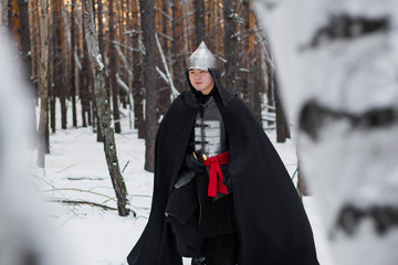 Fototapeta na wymiar Medieval warrior in armor, helmet, black cloak with a saber in his hands in the winter in the forest.