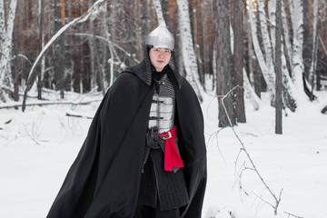 Medieval warrior in armor, helmet, black cloak with a saber in his hands in the winter in the forest.