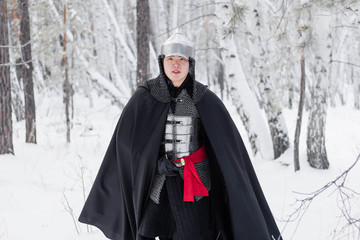Fototapeta premium Medieval warrior in armor, helmet, black cloak with a saber in his hands in the winter in the forest.