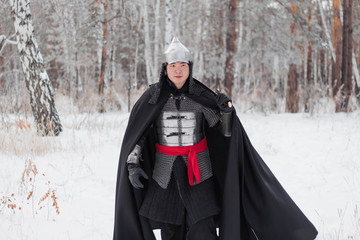 Medieval warrior in armor, helmet, black cloak with a saber in his hands in the winter in the forest.