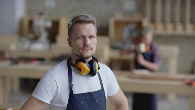 Waist up video portrait of calm middle aged carpenter looking at camera posing with his hands on hips  in carpentry shop while his defocused son learning to woodwork in background, copy space around