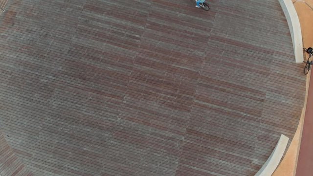 In the background, a girl rides a Bicycle on a round stadium, top view. She's wearing a blue jacket and jeans.