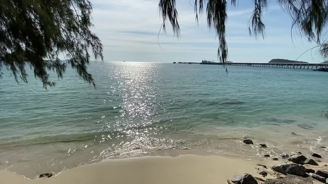 The Sea Is Wide During The Day Time, Not Very High Wave To Reach The Coast, Sand And The Rock Not Too Strong. It's A Picture Frame Consisting Of Pine Trees, Sunlight Reaching The Wave, Clear Blue Sky