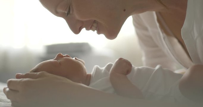 Mother and her newborn baby girl kissing, talking to her in bright bedroom setting. 