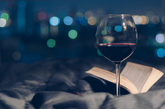 A glass of Red wine put on bed with book and colorful bokeh from city lights background.