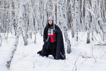 Medieval warrior in chain mail, helmet, black cloak with a saber in his hands against the background of a winter birch forest.
