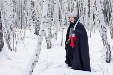 Medieval warrior in chain mail, helmet, black cloak with a saber in his hands against the background of a winter birch forest.