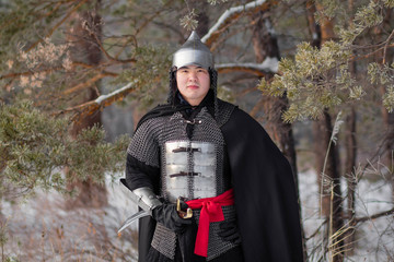    Portrait of a medieval warrior in armor, a helmet and a black cloak with a saber in his hands against the background of a winter forest an