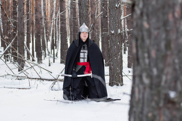 A medieval warrior in chain mail armor, a helmet and a black cloak with a saber in his hands, stands aside to a tree. Against the background of winter forest and snow.