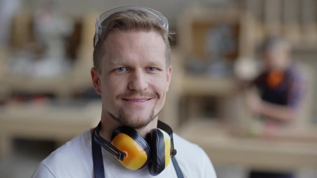 Head and shoulders portrait of middle aged carpenter looking at camera and smiling in carpentry shop with protective earmuffs around his neck while smb working in background, copy space to right
