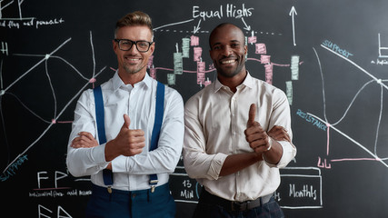 Financial best ideas pay off. Diverse employees, traders standing near blackboard full of charts and looking at camera, while discussing the strategy of work. Horizontal shot