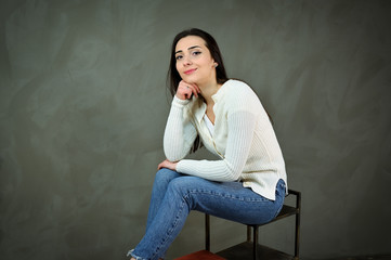 Portrait of a pretty young brunette woman in a white sweater and blue jeans sits on a stand. The concept of a cute girl on a gray background. Smiling, showing hands with emotions.