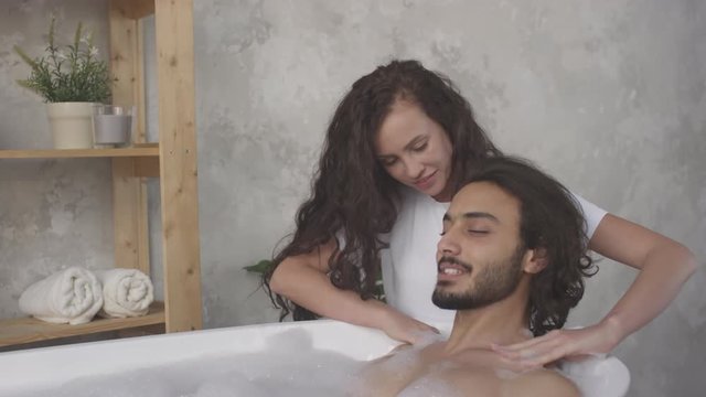 Beautiful brunette woman smiling and massaging shoulders of her boyfriend while he lying in modern freestanding bath