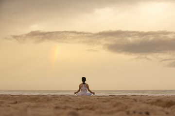 Yoga lotus pose meditation practice of young Asian female on an beach with rainbow background.